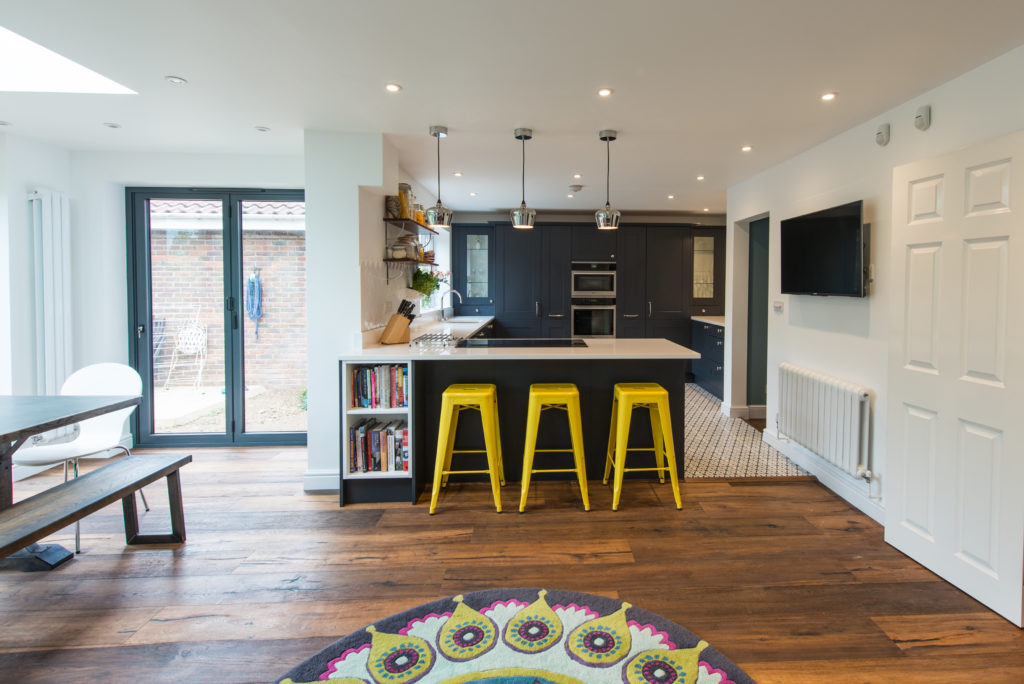 Image of an open-plan kitchen design with white walls, dark blue kitchen units, bright yellow kitchen accessories, and brown wooden flooring.