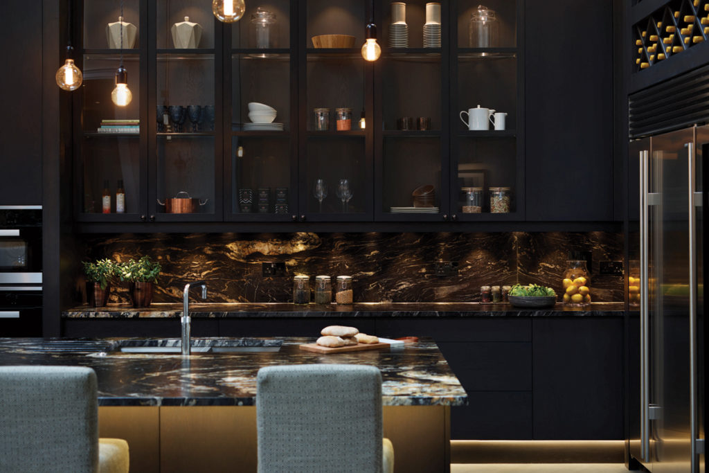 A decadent kitchen island with dark marble countertop, seating and ambient lighting.